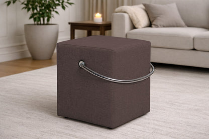 Brauner Polsterhocker mit Griff im modernen Wohnzimmer, leichter Sitzhocker auf hellem Teppich vor Sofa und Beistelltisch.– Handgefertigte Polsterhocker